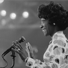 Photograph shows Shirley Chisholm smiling at microphone.