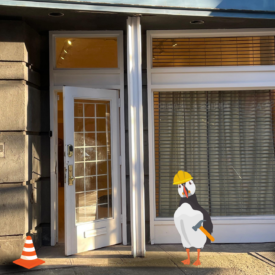 A photo of our new storefront. It is a large white framed window and door. In the foreground an illustrated puffin is wearing a hard hat