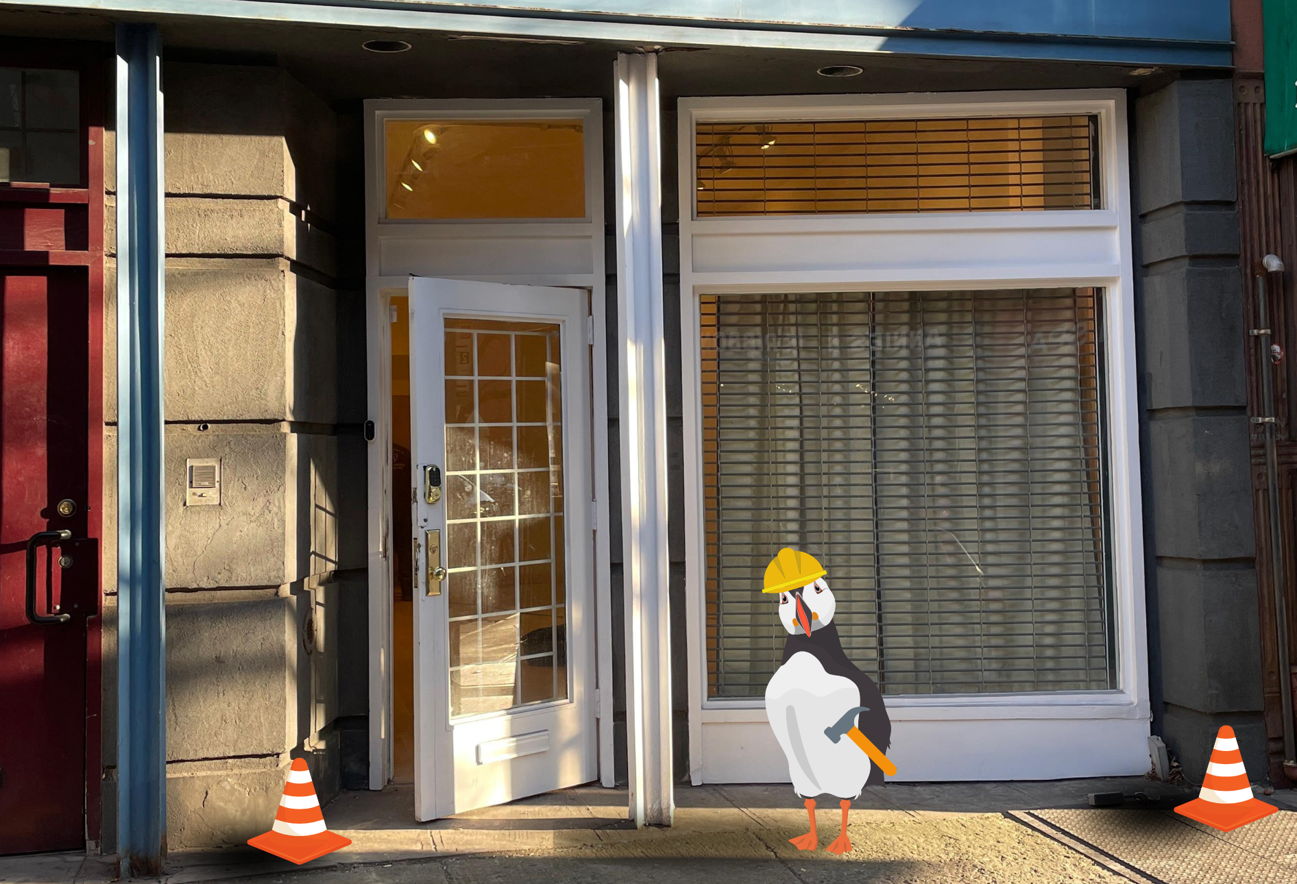 A photo of our new storefront. It is a large white framed window and door. In the foreground an illustrated puffin is wearing a hard hat